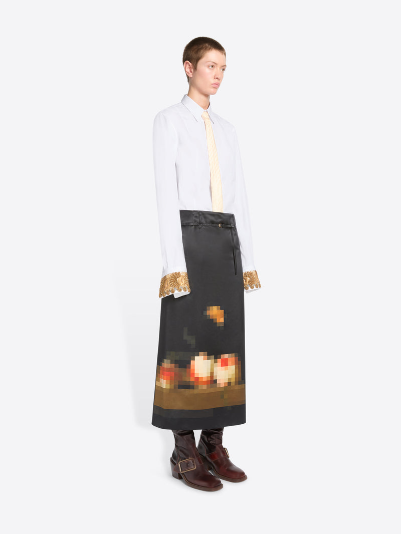 A model stands against a light gray background wearing a white long-sleeve shirt with gold lace cuffs, a cream tie, and a black skirt featuring a pixel-art flame graphic near the hem, paired with dark brown boots.