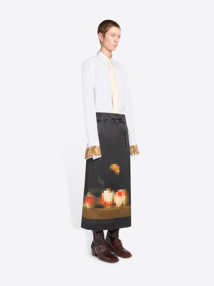 A model stands against a light gray background wearing a white long-sleeve shirt with gold lace cuffs, a cream tie, and a black skirt featuring a pixel-art flame graphic near the hem, paired with dark brown boots.