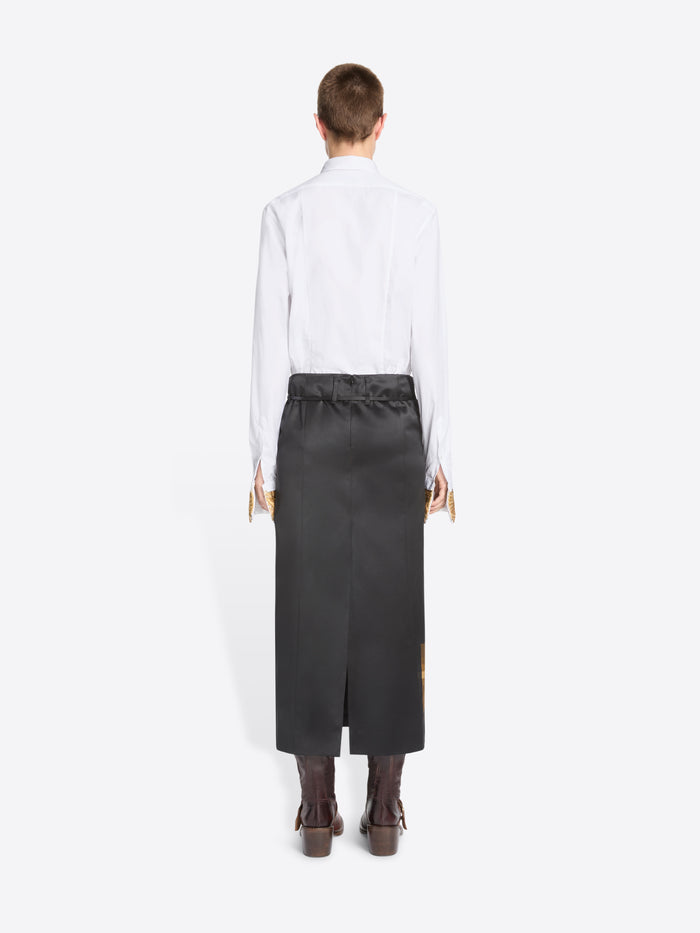 Back view of a model standing against a light gray background wearing a white long-sleeve shirt with gold lace cuffs, a cream tie, and a black skirt featuring a pixel-art flame graphic near the hem, paired with dark brown boots.