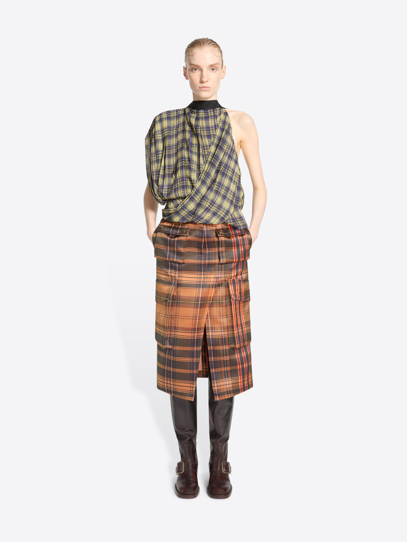 A model stands against a light gray background wearing a sleeveless asymmetrical plaid top in olive green and black, paired with a matching plaid wrap skirt in orange, brown, and black, finished with dark brown knee-high boots.