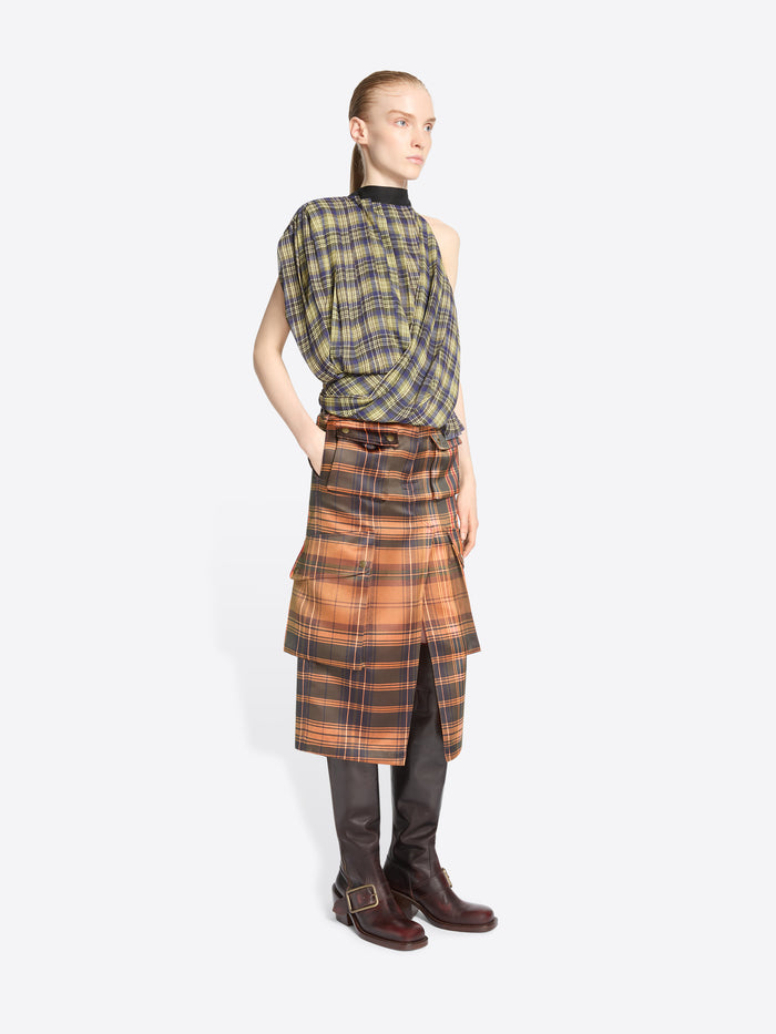 A model stands against a light gray background wearing a sleeveless asymmetrical plaid top in olive green and black, paired with a matching plaid wrap skirt in orange, brown, and black, finished with dark brown knee-high boots.