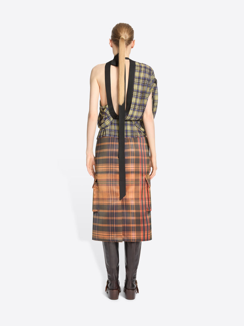 Back view of a model standing against a light gray background wearing a sleeveless asymmetrical plaid top in olive green and black, paired with a matching plaid wrap skirt in orange, brown, and black, finished with dark brown knee-high boots.