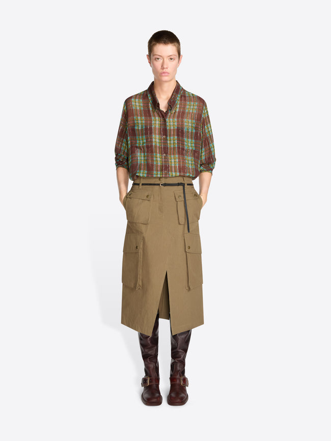A model stands against a light gray studio background wearing a green and red plaid button-up shirt tucked into a tan knee-length utility skirt with a black belt, paired with dark brown boots.