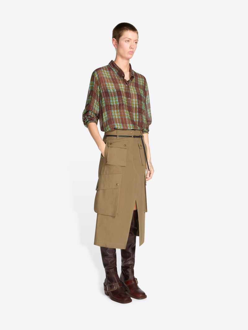 A model stands against a light gray studio background wearing a green and red plaid button-up shirt tucked into a tan knee-length utility skirt with a black belt, paired with dark brown boots.