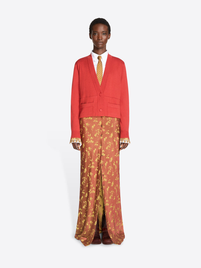 A model stands against a light gray studio background wearing a bright red cropped cardigan over a white collared shirt and a gold patterned tie, paired with rust-orange patterned skirt and dark brown shoes.
