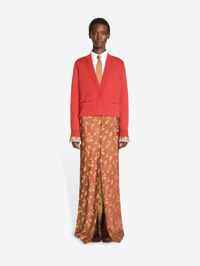 A model stands against a light gray studio background wearing a bright red cropped cardigan over a white collared shirt and a gold patterned tie, paired with rust-orange patterned skirt and dark brown shoes.