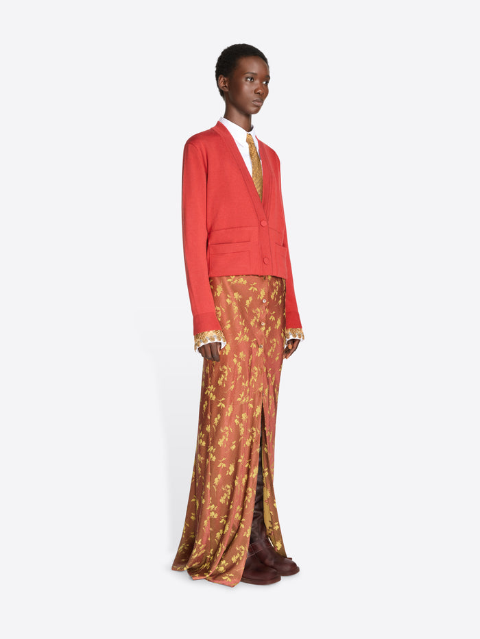 A model stands against a light gray studio background wearing a bright red cropped cardigan over a white collared shirt and a gold patterned tie, paired with rust-orange patterned skirt and dark brown shoes.