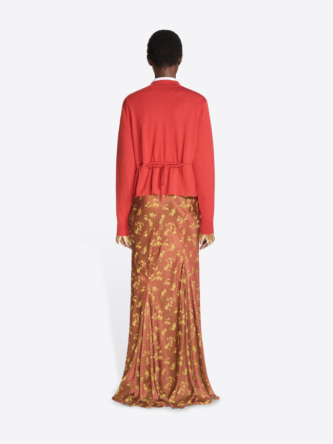Back view of a model standing against a light gray studio background wearing a bright red cropped cardigan over a white collared shirt and a gold patterned tie, paired with rust-orange patterned skirt and dark brown shoes.