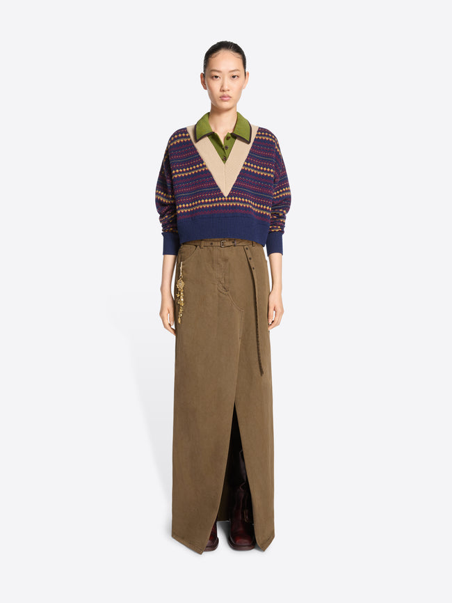 A model stands against a light gray background wearing a long brown skirt with a front slit and gold-tone chain detailing on the pocket, paired with a patterned knit sweater.