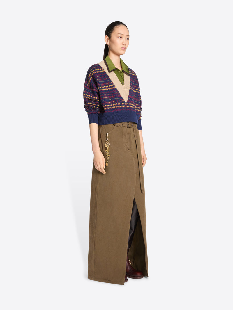 A model stands against a light gray background wearing a long brown skirt with a front slit and gold-tone chain detailing on the pocket, paired with a patterned knit sweater.