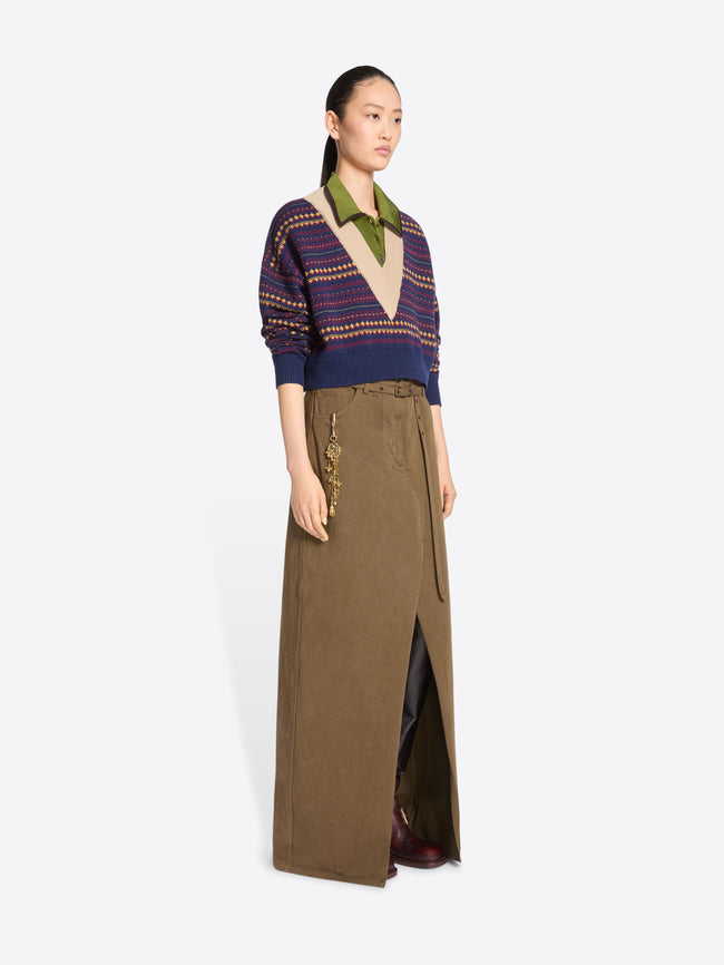 A model stands against a light gray background wearing a long brown skirt with a front slit and gold-tone chain detailing on the pocket, paired with a patterned knit sweater.