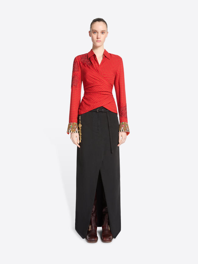 A model stands against a light gray background wearing a red long-sleeve wrap-style top with gold cuff embellishments and a black maxi skirt with a front slit.