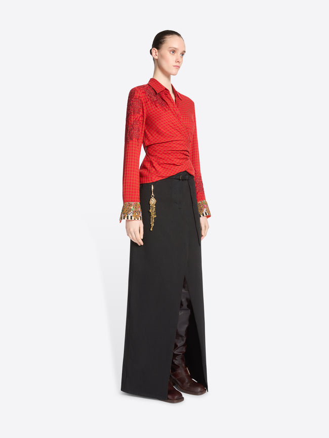 A model stands against a light gray background wearing a red long-sleeve wrap-style top with gold cuff embellishments and a black maxi skirt with a front slit.