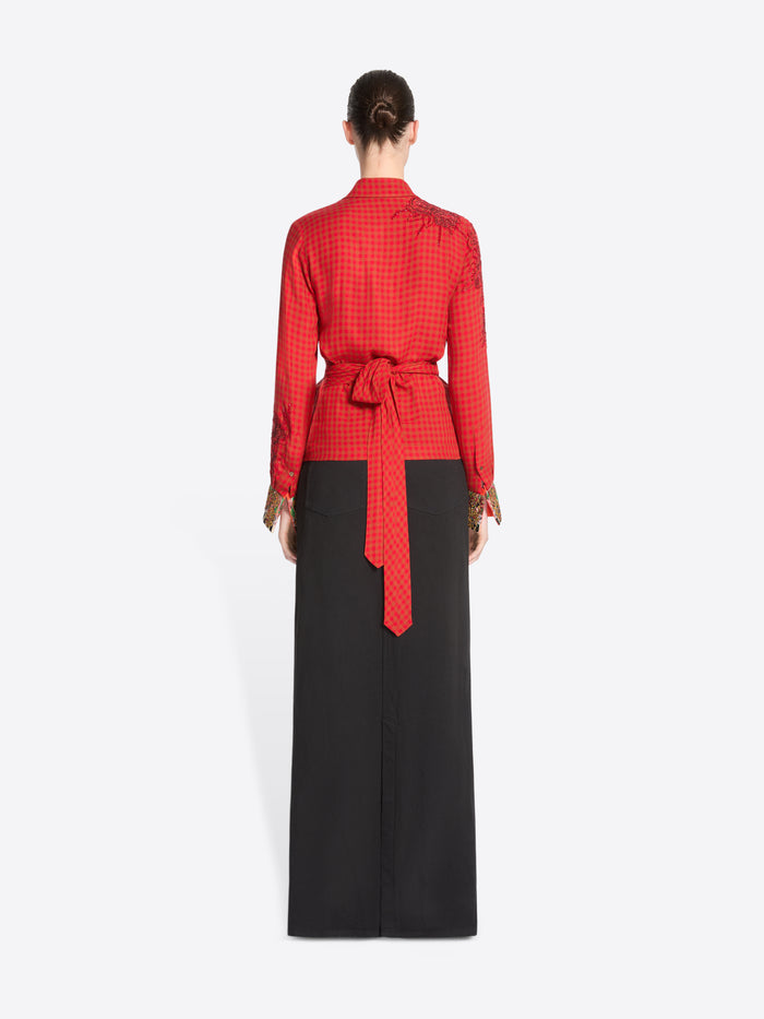 Back view of a model standing against a light gray background wearing a red long-sleeve wrap-style top with gold cuff embellishments and a black maxi skirt with a front slit.