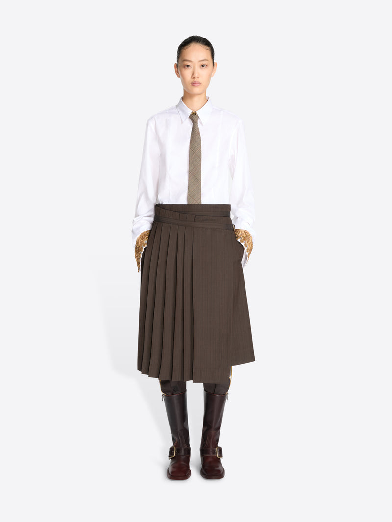 A model stands against a light gray background wearing a white long-sleeve button-up shirt, a taupe tie, a dark brown pleated midi skirt, and dark brown knee-high boots.