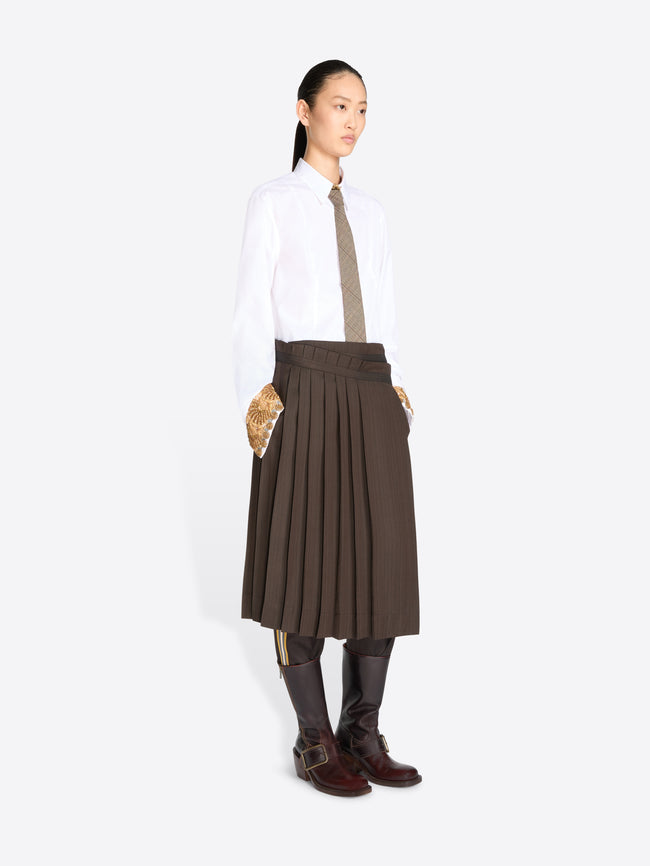 A model stands against a light gray background wearing a white long-sleeve button-up shirt, a taupe tie, a dark brown pleated midi skirt, and dark brown knee-high boots.
