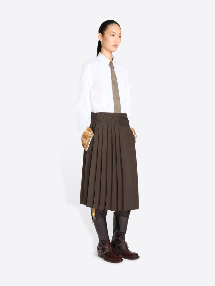 A model stands against a light gray background wearing a white long-sleeve button-up shirt, a taupe tie, a dark brown pleated midi skirt, and dark brown knee-high boots.
