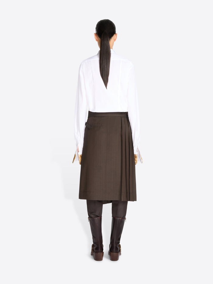 Back view of a model standing against a light gray background wearing a white long-sleeve button-up shirt, a taupe tie, a dark brown pleated midi skirt, and dark brown knee-high boots.