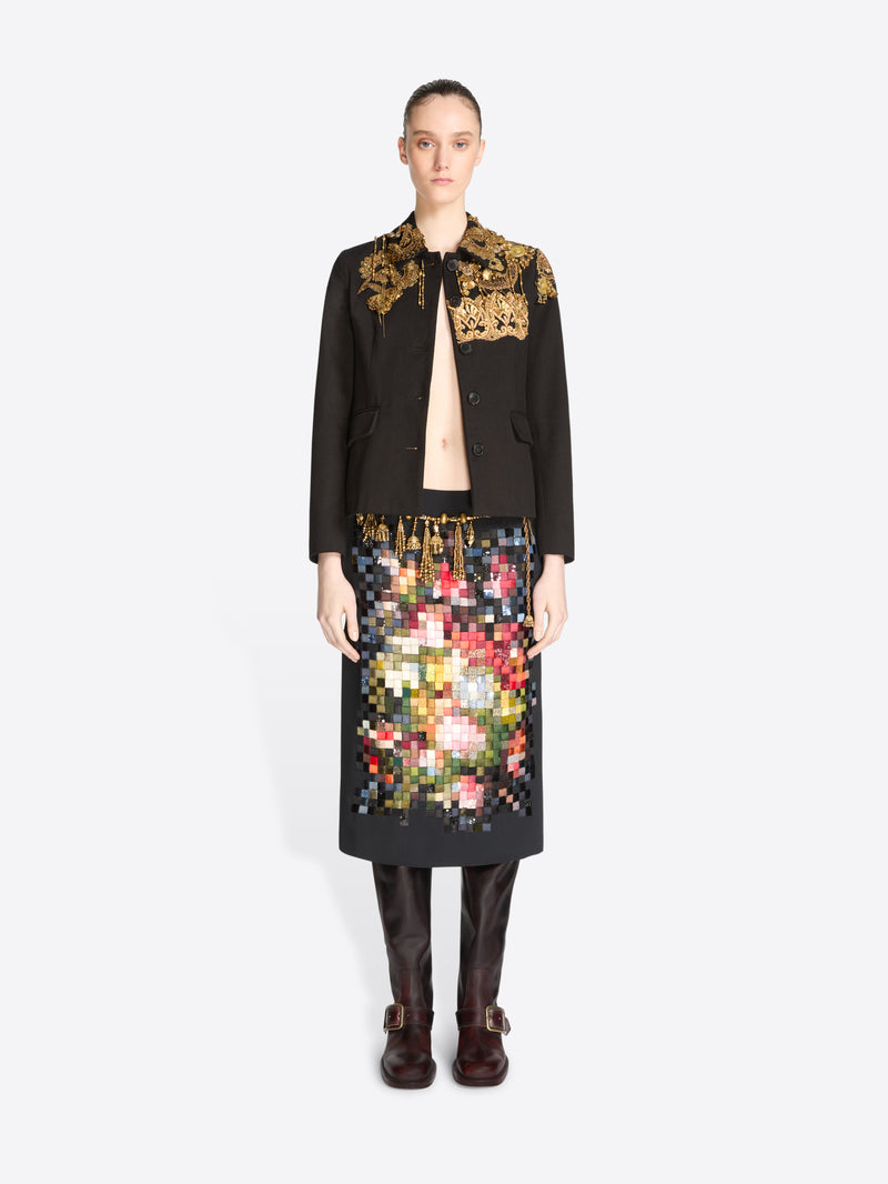 A model stands against a light gray background wearing a black tailored jacket with ornate gold embroidery on the shoulders and a black skirt covered in a colorful pixelated floral pattern, paired with dark brown knee-high boots.