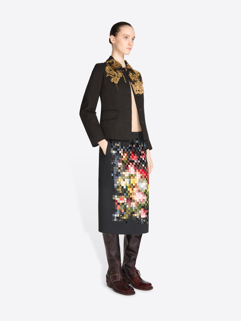A model stands against a light gray background wearing a black tailored jacket with ornate gold embroidery on the shoulders and a black skirt covered in a colorful pixelated floral pattern, paired with dark brown knee-high boots.