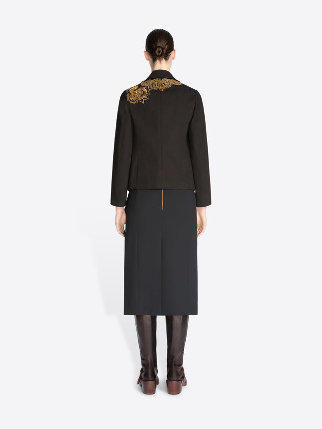 Back view of a model standing against a light gray background wearing a black tailored jacket with ornate gold embroidery on the shoulders and a black skirt, paired with dark brown knee-high boots.