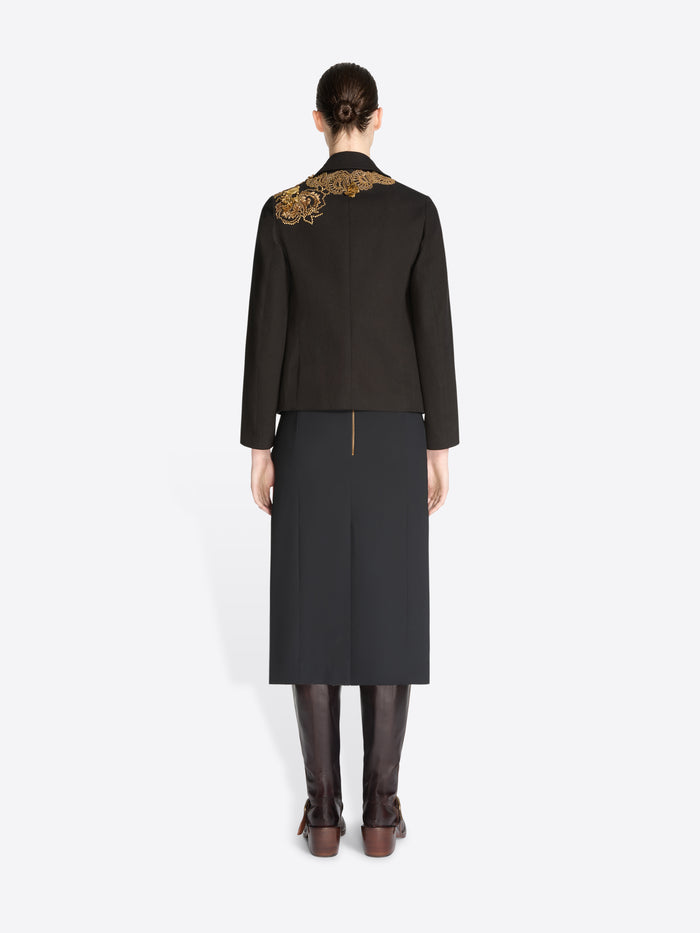 Back view of a model standing against a light gray background wearing a black tailored jacket with ornate gold embroidery on the shoulders and a black skirt, paired with dark brown knee-high boots.