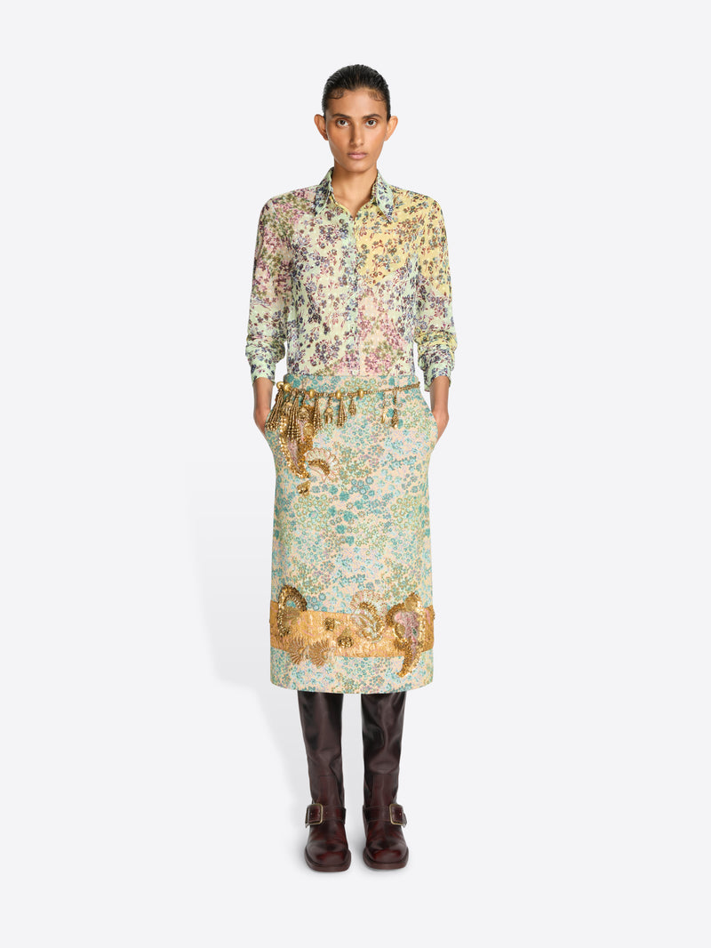 A model wearing a long-sleeved shirt with a dense multicolor floral and a floral skirt with paisley print in green, beige, and gold tones, paired with dark brown leather boots, against a light gray background.