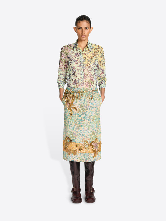 A model wearing a long-sleeved shirt with a dense multicolor floral and a floral skirt with paisley print in green, beige, and gold tones, paired with dark brown leather boots, against a light gray background.
