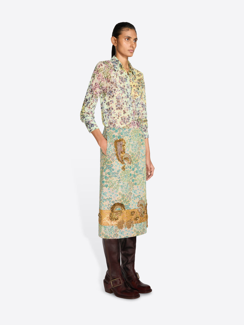 A model wearing a long-sleeved shirt with a dense multicolor floral and a floral skirt with paisley print in green, beige, and gold tones, paired with dark brown leather boots, against a light gray background.