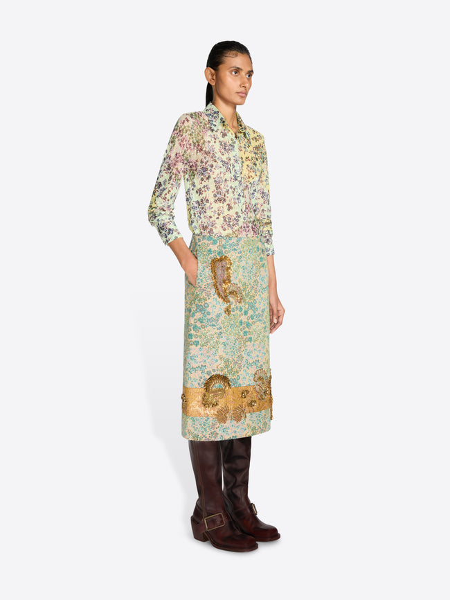 A model wearing a long-sleeved shirt with a dense multicolor floral and a floral skirt with paisley print in green, beige, and gold tones, paired with dark brown leather boots, against a light gray background.