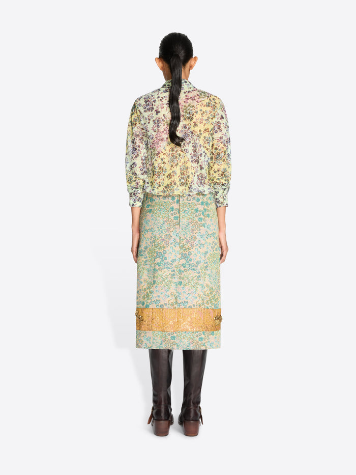 Back view of a model wearing a long-sleeved shirt with a dense multicolor floral and a floral skirt with paisley print in green, beige, and gold tones, paired with dark brown leather boots, against a light gray background.