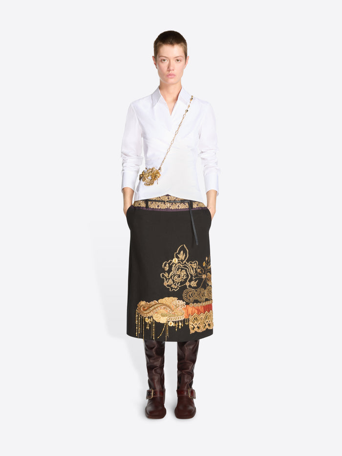 A model stands against a light gray background wearing a white long-sleeve shirt, a black midi skirt with ornate gold and red printed motifs, dark brown knee-high boots, and a small gold crossbody bag.