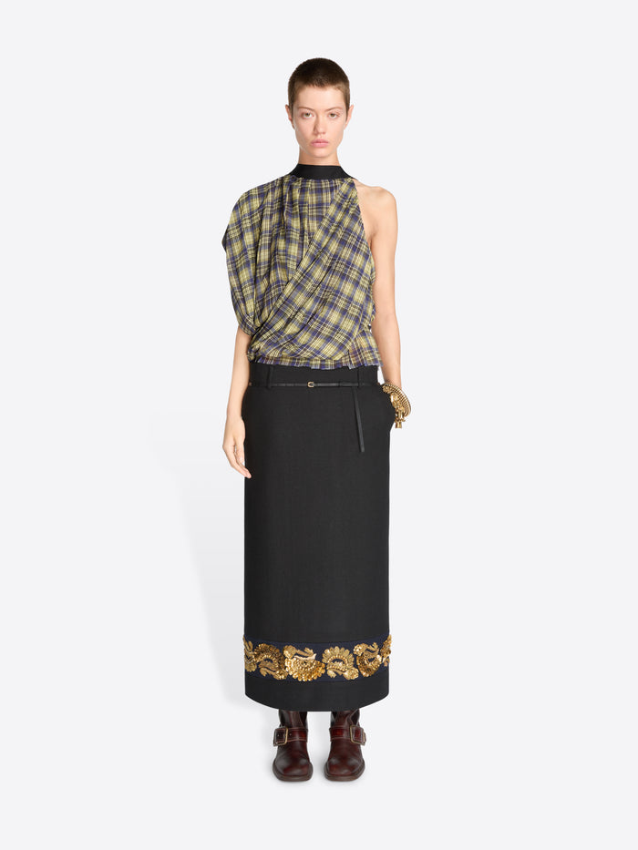 A model wearing a sleeveless plaid top in muted green, yellow, and gray tones, paired with a long black skirt featuring ornate gold embroidery near the hem, and dark brown boots, against a light gray background.