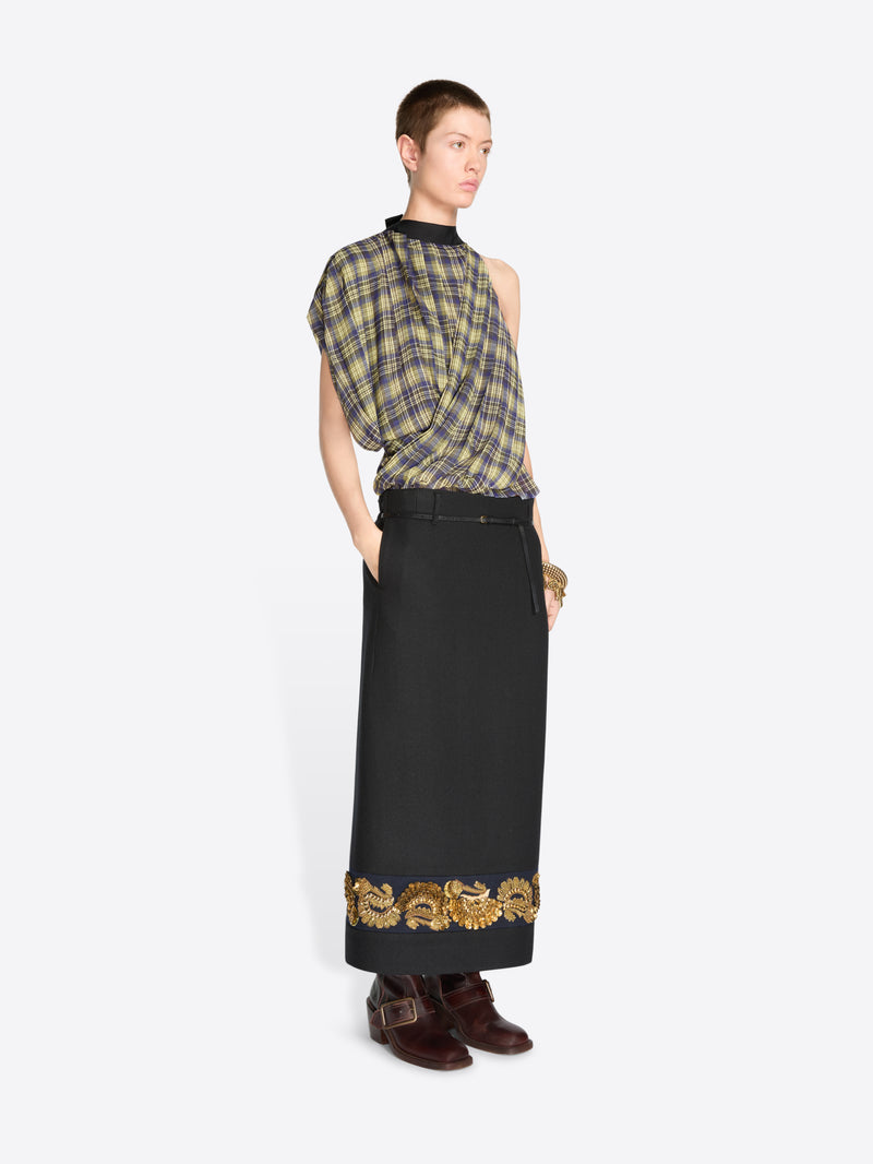 A model wearing a sleeveless plaid top in muted green, yellow, and gray tones, paired with a long black skirt featuring ornate gold embroidery near the hem, and dark brown boots, against a light gray background.