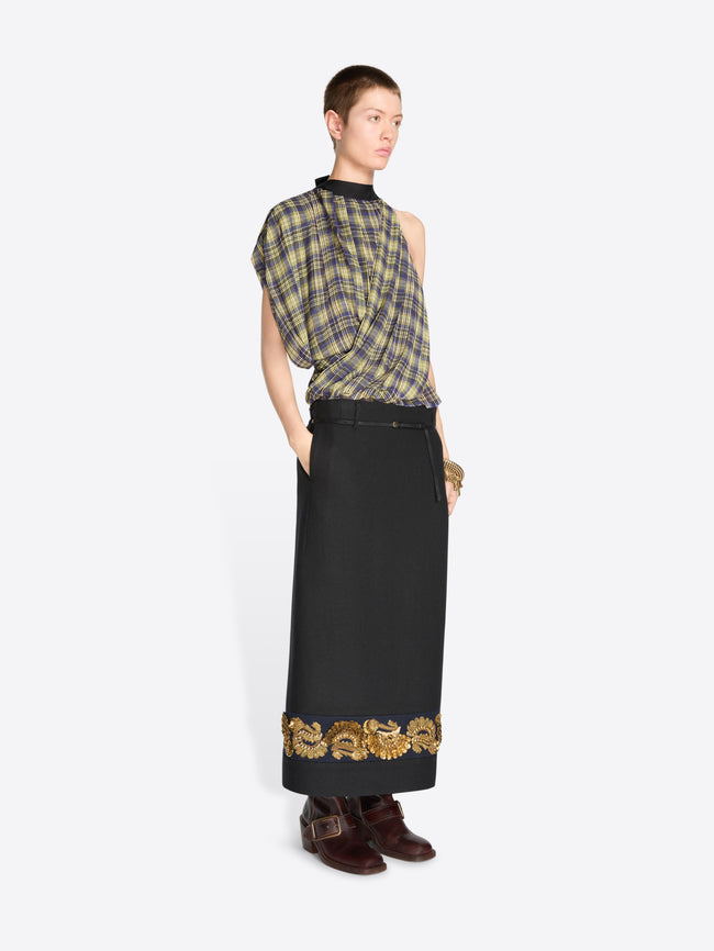 A model wearing a sleeveless plaid top in muted green, yellow, and gray tones, paired with a long black skirt featuring ornate gold embroidery near the hem, and dark brown boots, against a light gray background.