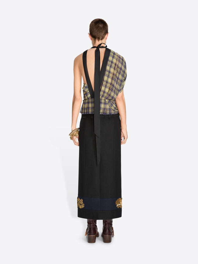 Back view of a model wearing a sleeveless plaid top in muted green, yellow, and gray tones, paired with a long black skirt featuring ornate gold embroidery near the hem, and dark brown boots, against a light gray background.