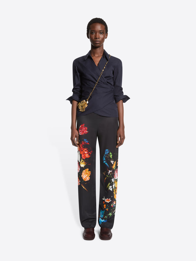 A model stands against a light gray background wearing a dark navy wrap-style blouse with rolled sleeves, black wide-leg trousers with colorful floral prints, and dark brown shoes, paired with a small gold crossbody bag. 