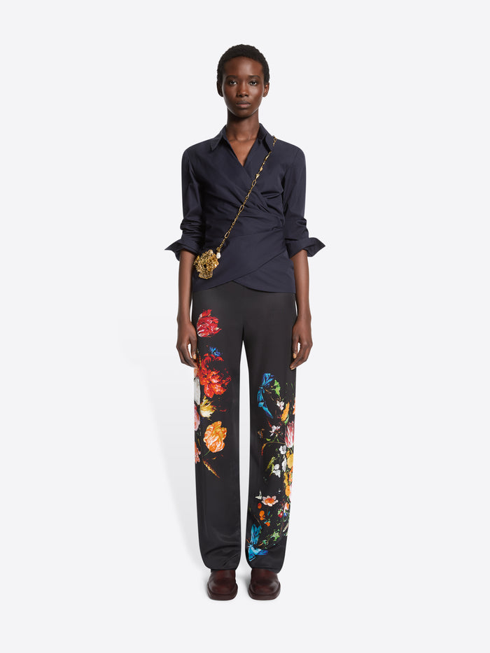 A model stands against a light gray background wearing a dark navy wrap-style blouse with rolled sleeves, black wide-leg trousers with colorful floral prints, and dark brown shoes, paired with a small gold crossbody bag. 