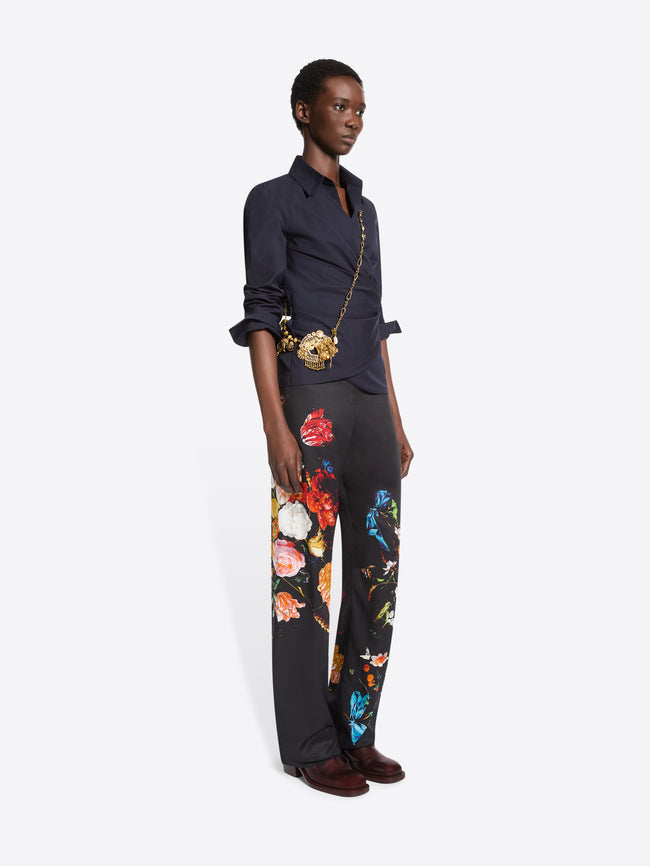 A model stands against a light gray background wearing a dark navy wrap-style blouse with rolled sleeves, black wide-leg trousers with colorful floral prints, and dark brown shoes, paired with a small gold crossbody bag.