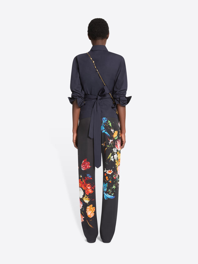 Back view of a model standing against a light gray background wearing a dark navy wrap-style blouse with rolled sleeves, black wide-leg trousers with colorful floral prints, and dark brown shoes, paired with a small gold crossbody bag. 