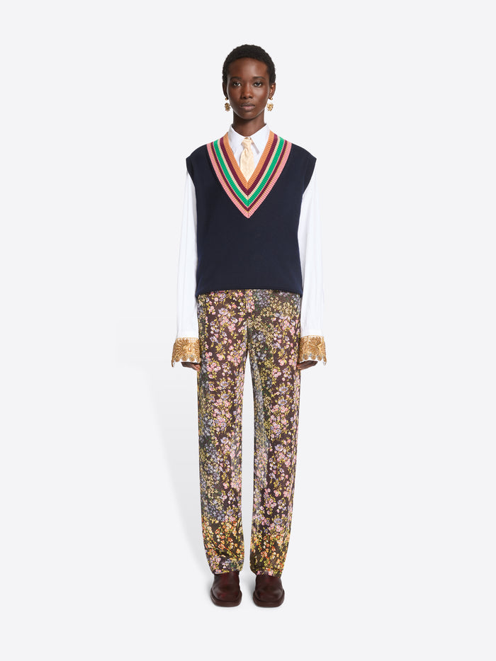 A model stands against a light gray background wearing a navy knit sweater vest with a colorful striped V-neck over a white collared shirt, paired with patterned trousers featuring a dense floral print and dark brown shoes.