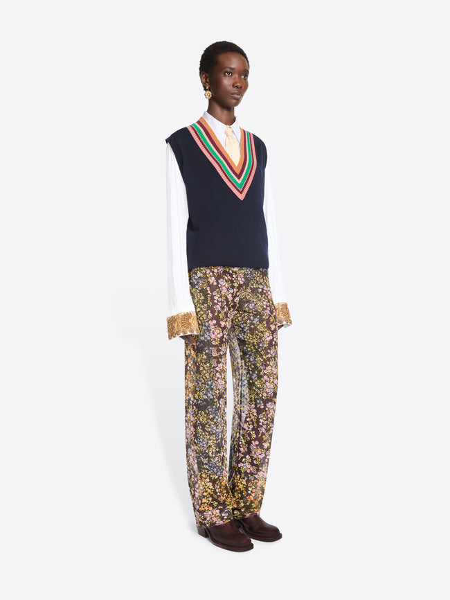 A model stands against a light gray background wearing a navy knit sweater vest with a colorful striped V-neck over a white collared shirt, paired with patterned trousers featuring a dense floral print and dark brown shoes.