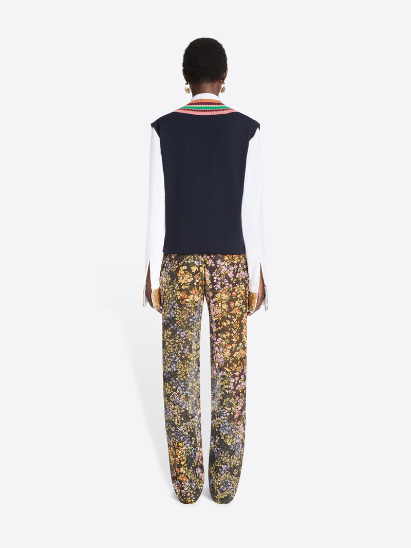 Back view of a model standing against a light gray background wearing a navy knit sweater vest with a colorful striped V-neck over a white collared shirt, paired with patterned trousers featuring a dense floral print and dark brown shoes.