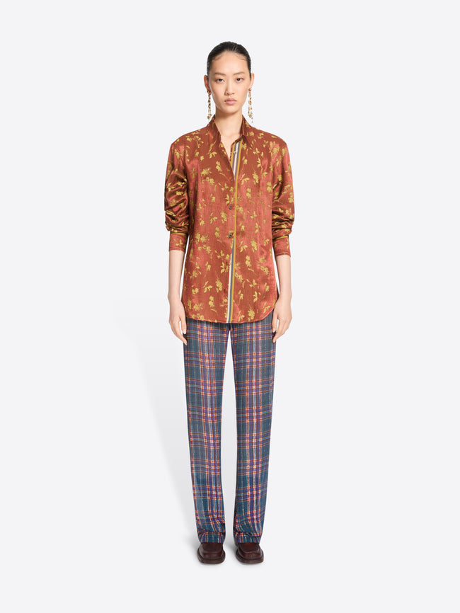A model stands against a light gray background wearing a rust-orange patterned button-up shirt with a front zipper and blue-red plaid trousers.