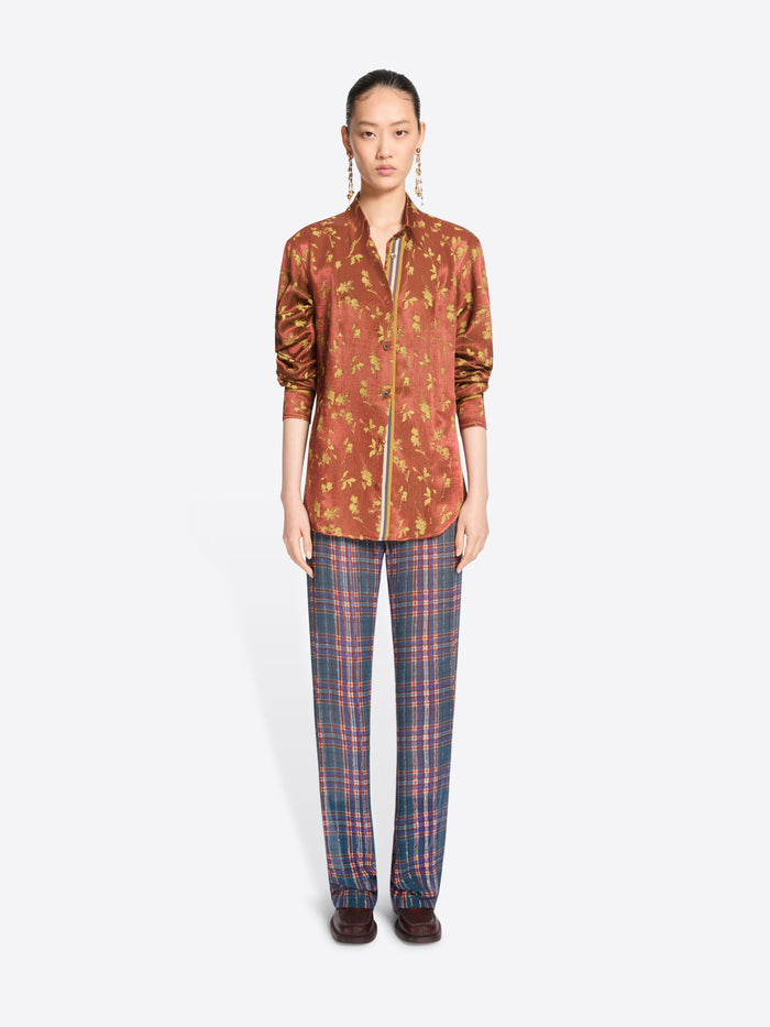 A model stands against a light gray background wearing a rust-orange patterned button-up shirt with a front zipper and blue-red plaid trousers.