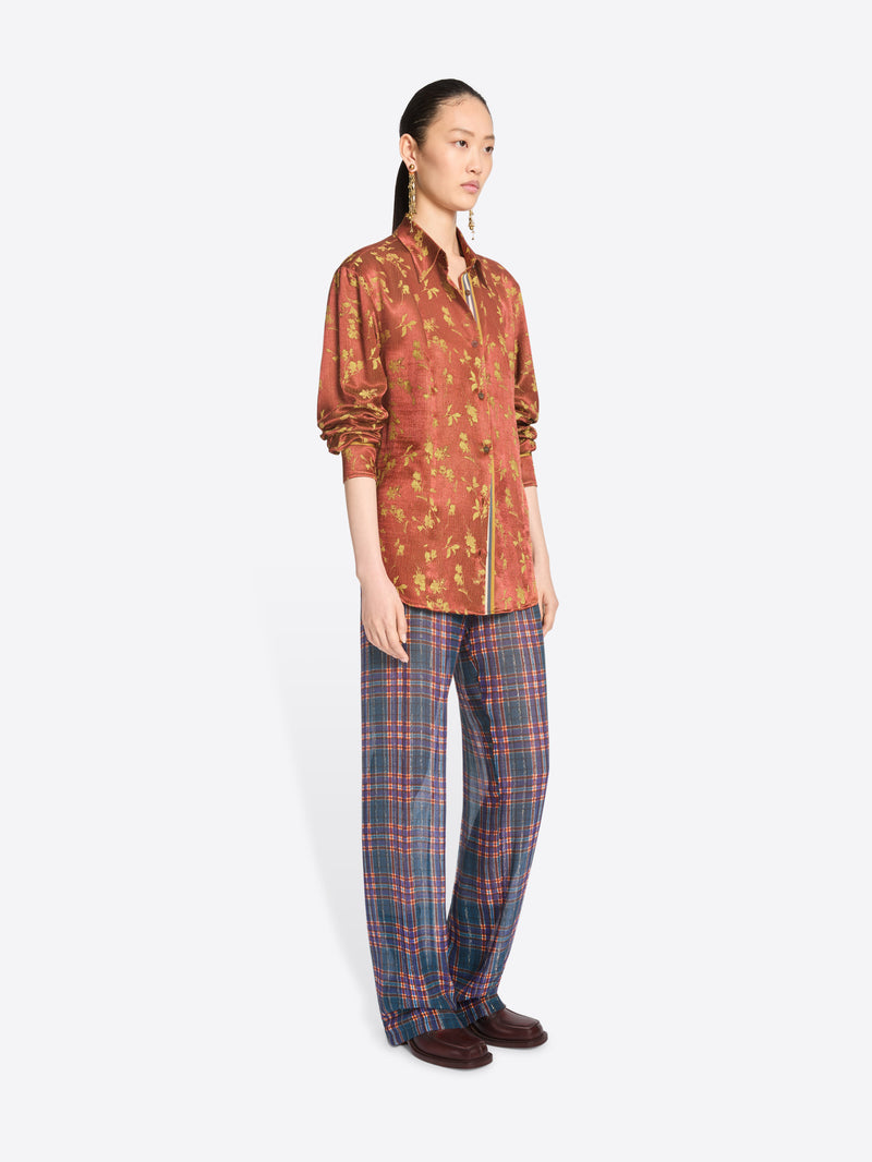 A model stands against a light gray background wearing a rust-orange patterned button-up shirt with a front zipper and blue-red plaid trousers.