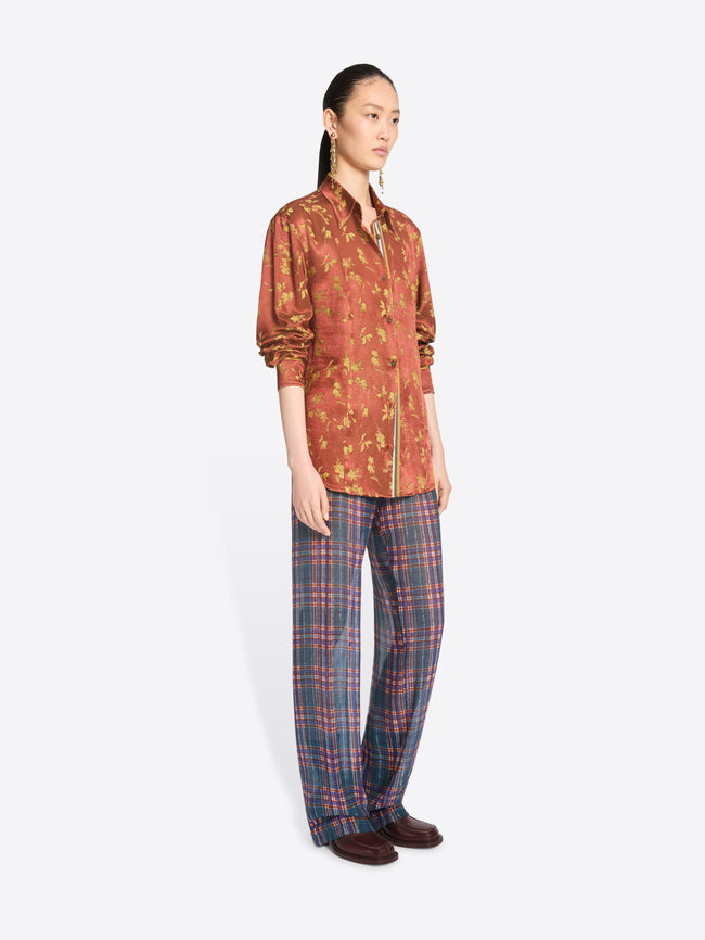 A model stands against a light gray background wearing a rust-orange patterned button-up shirt with a front zipper and blue-red plaid trousers.