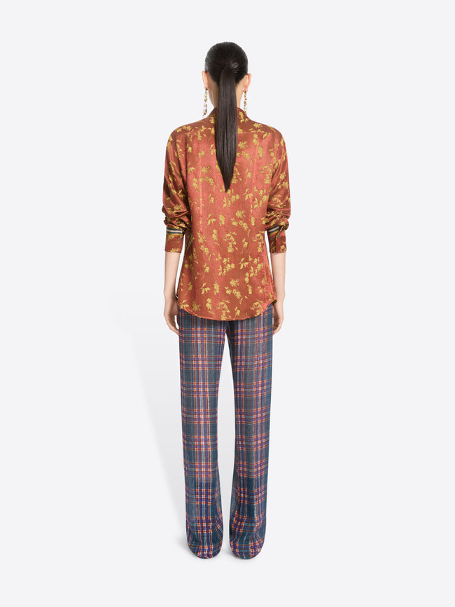 Back view of a model standing against a light gray background wearing a rust-orange patterned button-up shirt with a front zipper and blue-red plaid trousers.