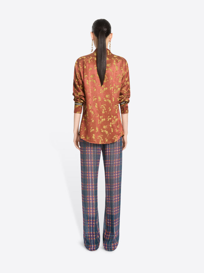 Back view of a model standing against a light gray background wearing a rust-orange patterned button-up shirt with a front zipper and blue-red plaid trousers.