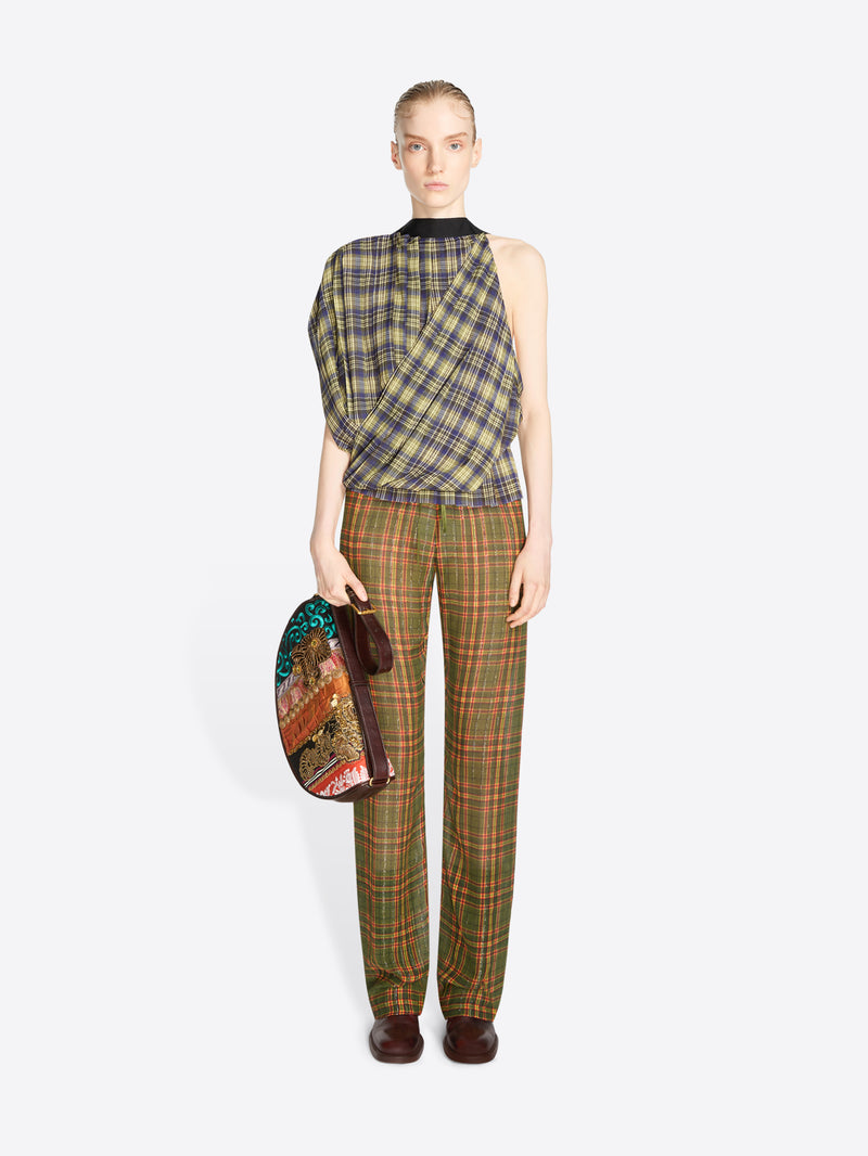 A model stands against a light gray background wearing a sleeveless asymmetrical plaid top and matching plaid trousers in olive green, brown, and beige tones, holding a patterned handbag.